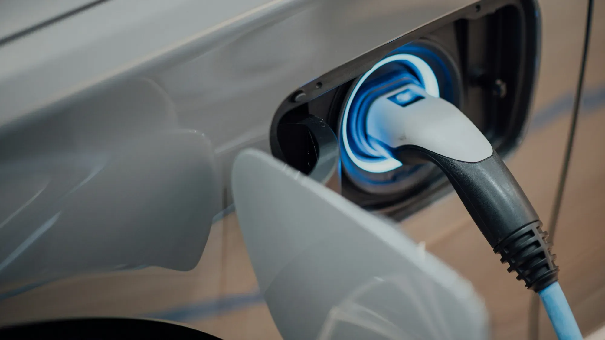Cable de carga con conector iluminado en azul, enchufado al puerto de un carro eléctrico gris