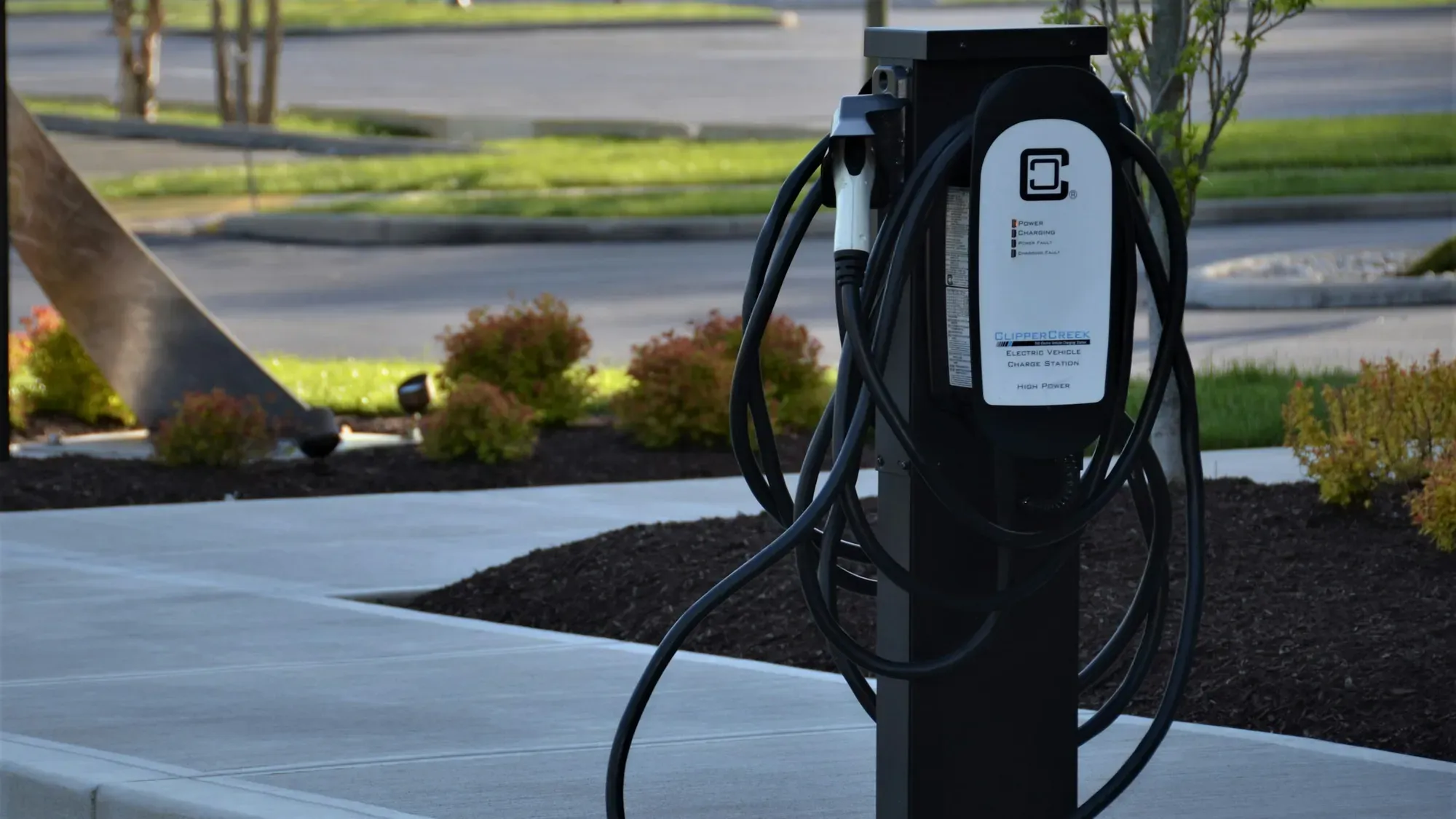 Cargador público para carros eléctricos en un pedestal negro, con su cable enrollado, junto a un andén
